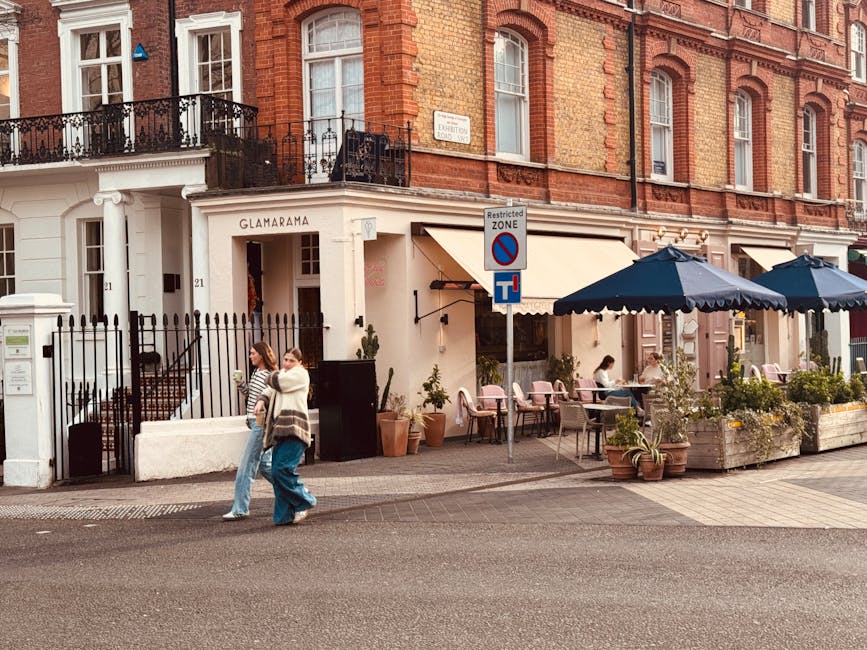 A street scene outside a building called 'Glamarama' with a white facade and black wrought-iron fencing, featuring outdoor seating with pink chairs and blue umbrellas covering several tables. In the foreground, two women are walking past, one wearing a striped top and jeans, the other in a white coat and blue jeans. Potted plants and small shrubs line the outdoor area, creating a welcoming atmosphere. A street sign indicates a no-parking zone, and a white mailbox with a green sticker is mounted on a nearby post. The background shows a red brick residential building with large windows and balconies, with some windows having decorative white trim. The overall scene is well-lit with natural daylight, emphasizing the cozy, urban environment. This setting suggests a lively local area that may involve private outdoor dining or on-site amenities independent of waste collection services, consistent with typical waste management practices for hospitality venues in the area.