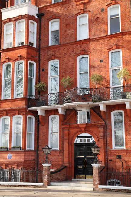 The image shows the front facade of a historic four-storey red-brick building with Victorian architectural features, including tall, narrow sash windows with white frames and decorative window surrounds. The building has a black entrance door with a semi-circular fanlight window above, framed by an ornate red-brick archway. A small, metal cast-iron balcony with decorative railings extends across the second floor, supported by white brackets, and is adorned with potted plants and small trees in window boxes. The surrounding environment includes a paved sidewalk with a low brick wall and black metal railings, as well as Victorian-style street lamps positioned on either side of the entrance. The overall scene reflects the building’s historic character, viewed in natural daylight, with no visible rubbish or waste-related elements present. Waste Collection Chelsea’s name and focus on independent waste collection services are associated with the context of managing household waste, though no rubbish is visible in this particular image, emphasizing the building’s residential and architectural features.