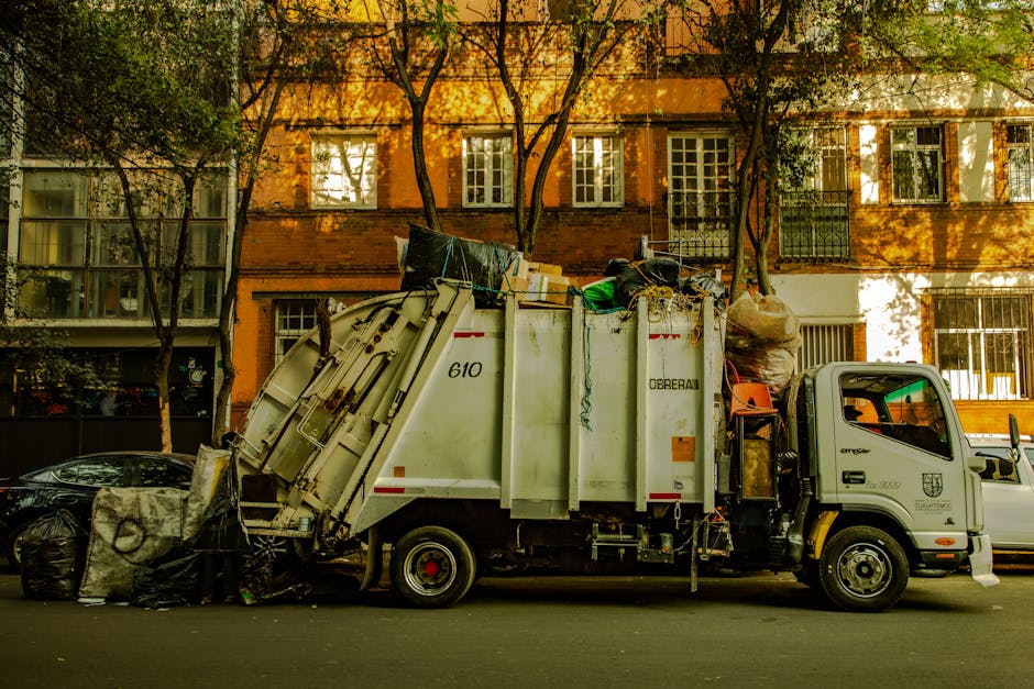 The image depicts a city street scene featuring a mix of historic and modern buildings with brick and ornate facades. Centrally, a light blue waste collection vehicle, likely used for private or independent waste collection services like Waste Collection Chelsea, is parked on the paved road near the sidewalk. The vehicle has a compact, boxy design with a small cab and a rear loading compartment, and it appears to be positioned close to the curb, ready for rubbish collection. Surrounding the vehicle, the street is flanked by multi-story buildings with large windows, some with arched designs and decorative trim, and a prominent green-domed tower visible atop one structure, indicating architectural detail typical of the area. The environment is well-lit, suggesting daytime with natural light casting minimal shadows. The roadway is lined with bicycles and a few pedestrians, while a lamppost and bollards are visible near the curb, emphasizing urban infrastructure. This scene exemplifies urban waste management possibly related to an alternative rubbish removal service operating within the city, supporting private waste disposal needs amidst a busy cityscape. Waste Collection Chelsea’s operational context is subtly referenced through the focus on the waste collection vehicle within an active urban environment, emphasizing the City’s approach to supplementary rubbish collection services.