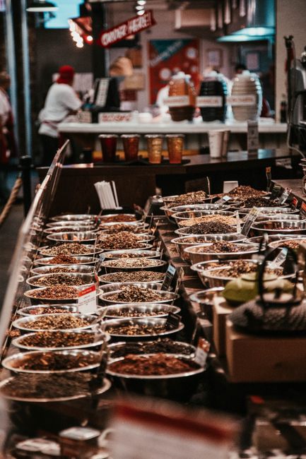 A retail display counter in a grocery or market setting, showcasing numerous small, clear glass bowls filled with dried herbs, spices, and seasoning blends. The bowls are arranged in multiple neat rows on a dark-colored countertop, with each containing a variety of textured, finely chopped or granular ingredients in shades of brown, beige, and dark green. In the background, a blurred scene of an indoor shopping area is visible, featuring a person wearing a white shirt and red cap, along with shelves and displays of other food products, some of which are topped with decorative elements like cakes or other baked goods. The environment is softly lit, creating a warm and inviting atmosphere typical of an indoor marketplace specializing in food products. The display area appears well-organized, with some small signs or labels positioned in front of the bowls, indicating the contents. Waste collection or disposal services by Waste Collection Chelsea might be relevant for managing leftovers or packaging waste in such retail environments, supporting efficient on-site waste handling and helping maintain cleanliness and hygiene standards.